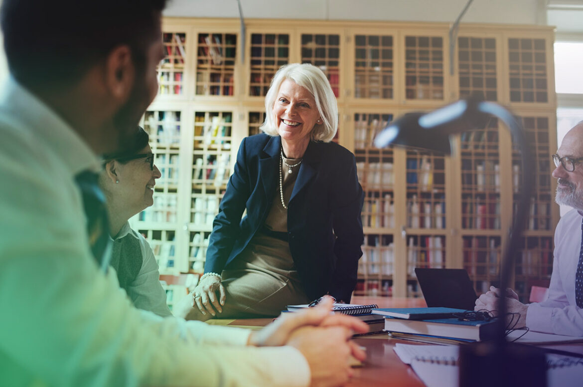 A law firm business owner is sitting on a table while she discusses with colleagues.